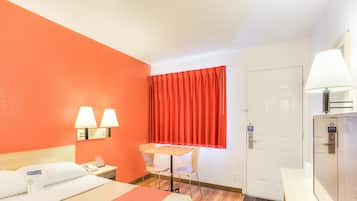 A hotel room with a bed, a desk, a chair, a window with red curtains, and a door.
