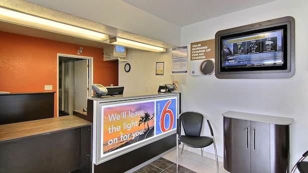 Zona de recepción de un hotel con una pantalla digital que muestra información meteorológica, un letrero que indica cargo por no tener acceso gratuito, y un mostrador con un cartel publicitario del hotel.