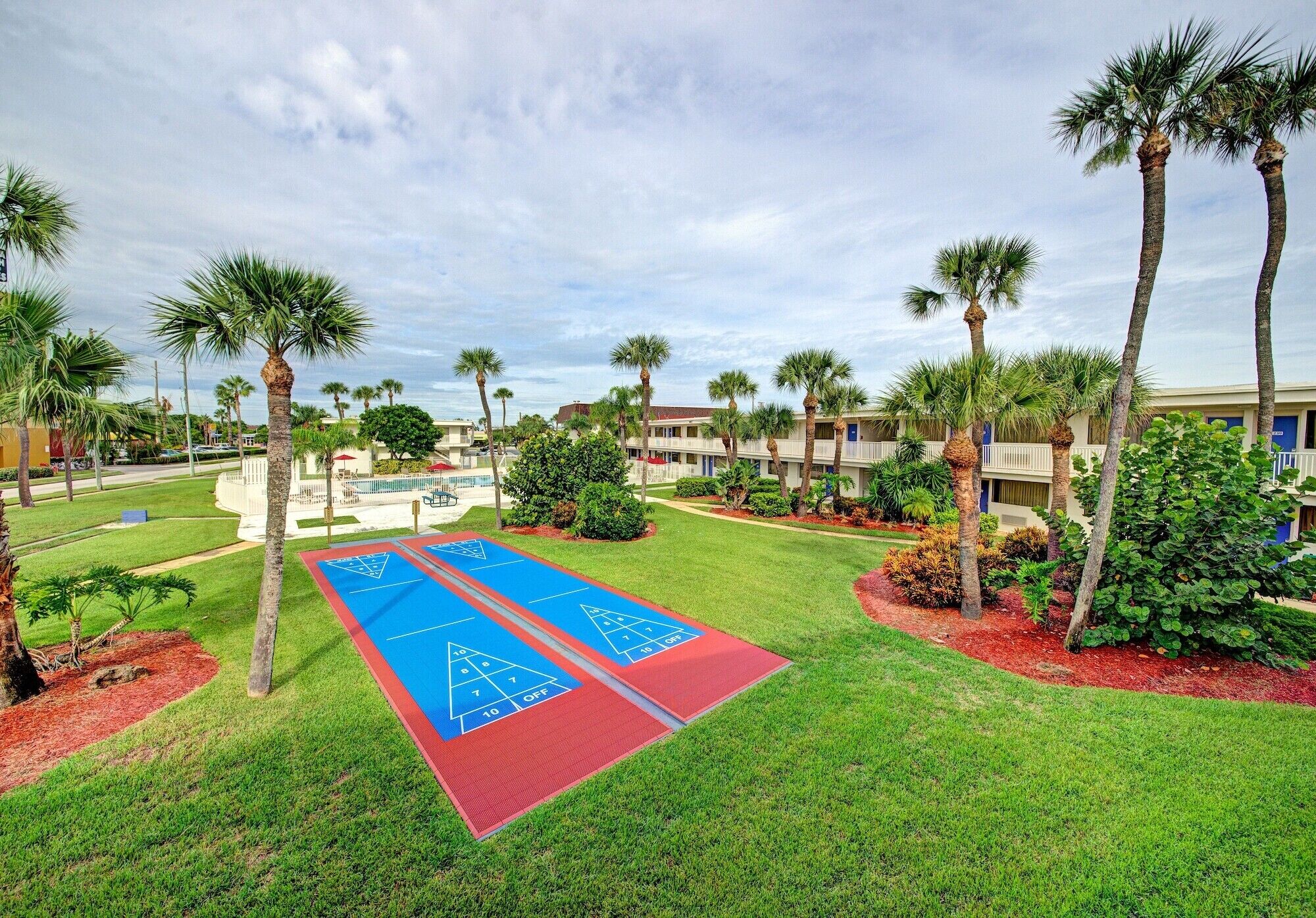 A courtyard with two shuffleboard courts, palm trees, and a building in the background.