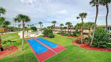 A courtyard with two shuffleboard courts, palm trees, and a building in the background.