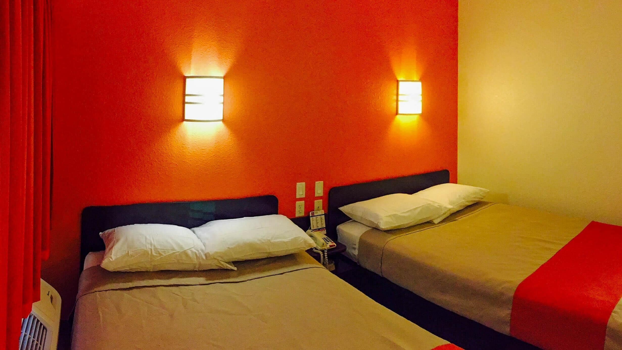 A hotel room with two beds, each with a pillow and a blanket. There are two wall-mounted light fixtures on the red wall.