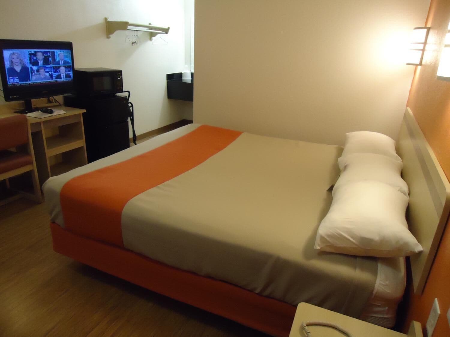 A hotel room with a bed, a desk with a computer, and a television displaying a news broadcast.
