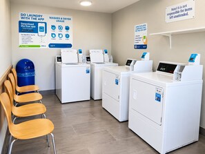 A laundry facility with four washing machines, a blue trash bin, and wooden chairs. - Motel 6 Amarillo, TX - West (Amarillo)