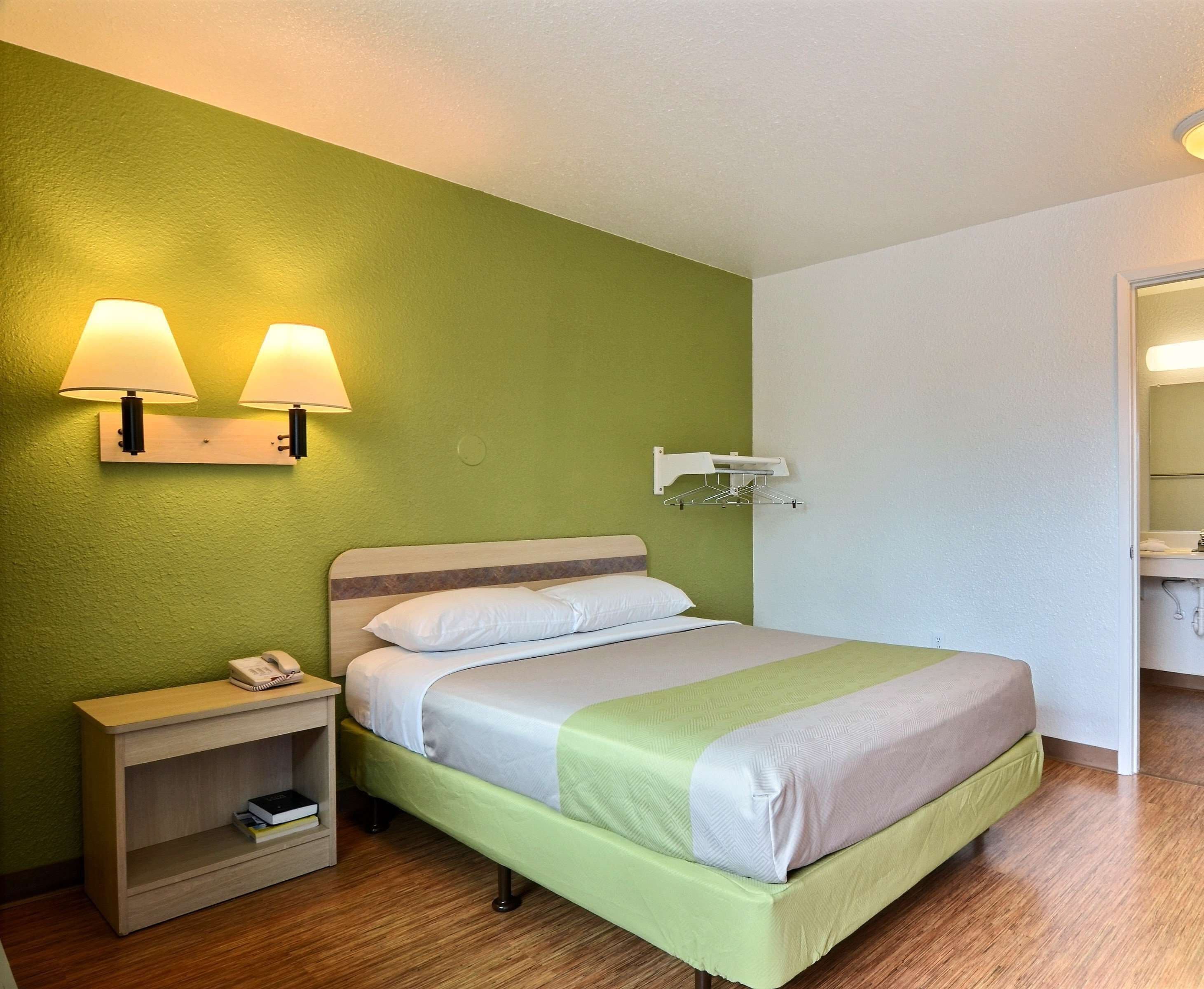 Quarto de hotel bem arrumado, com cama, mesa de cabeceira, abajur e um cabideiro.