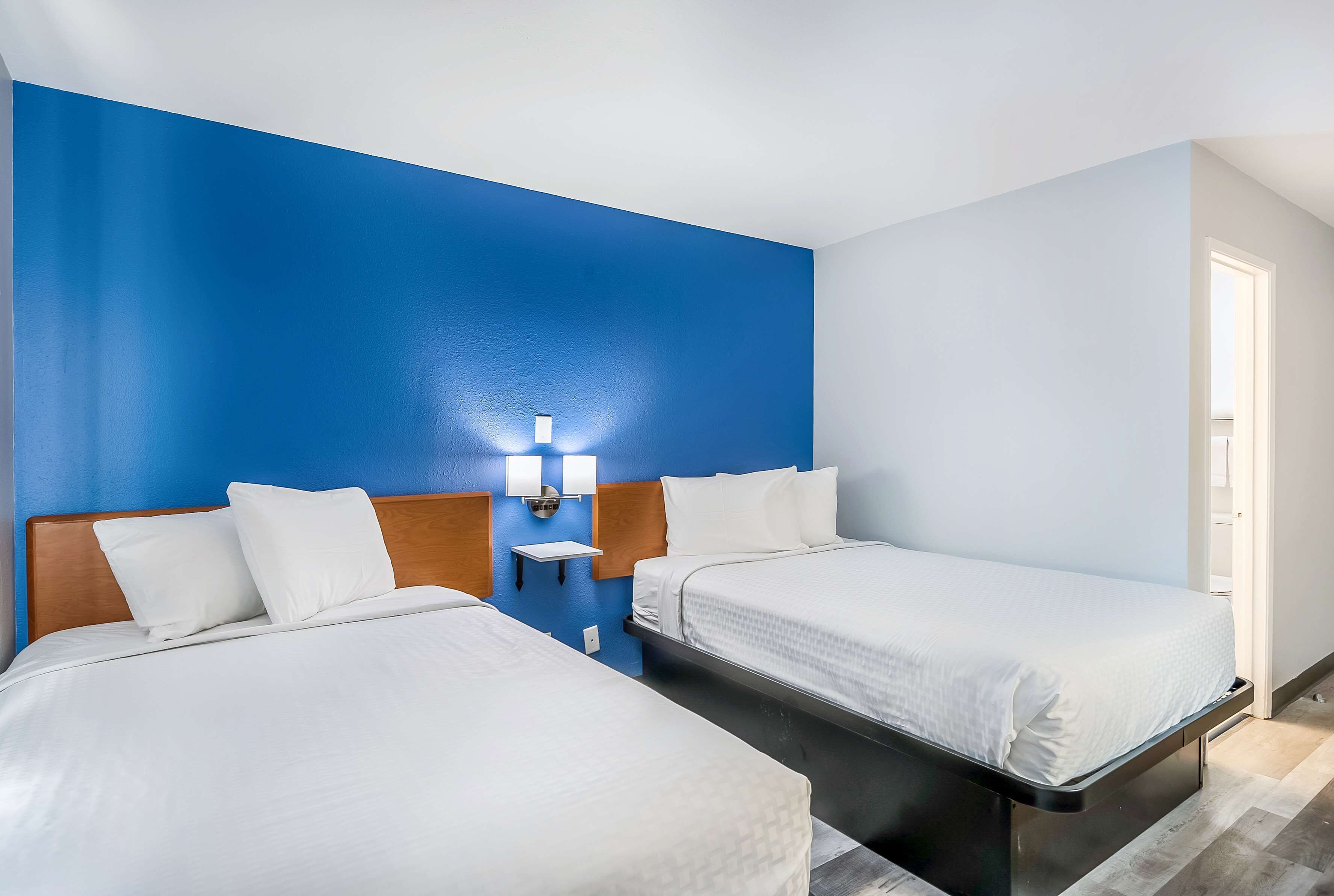 A hotel room with two beds, a blue accent wall, and a small bedside table with a lamp.