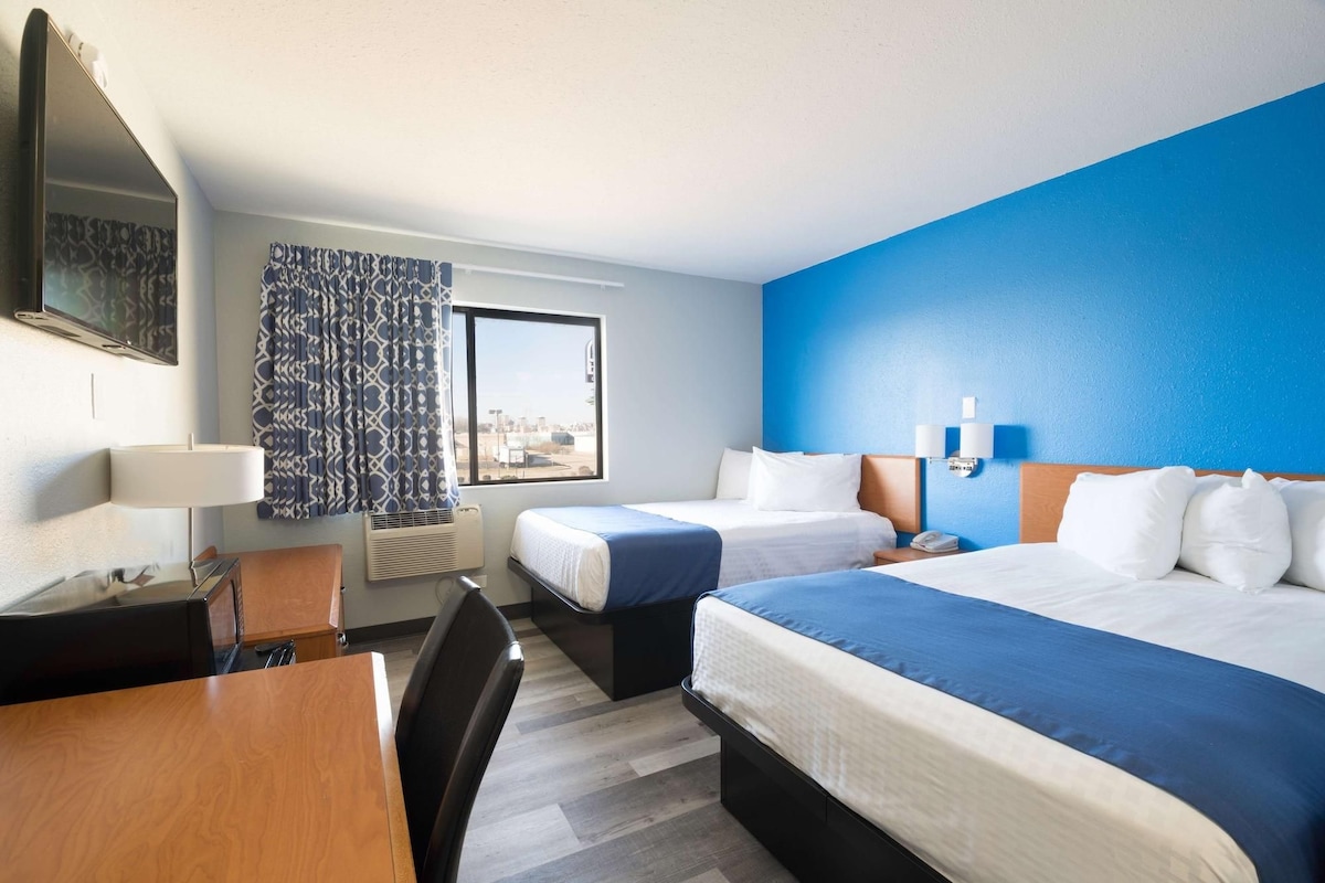 Ein Hotelzimmer mit zwei Betten, einem Schreibtisch, einem Stuhl, einer Schreibtischlampe, einem Fenster mit Vorhängen und einem an der Wand befestigten Fernseher.