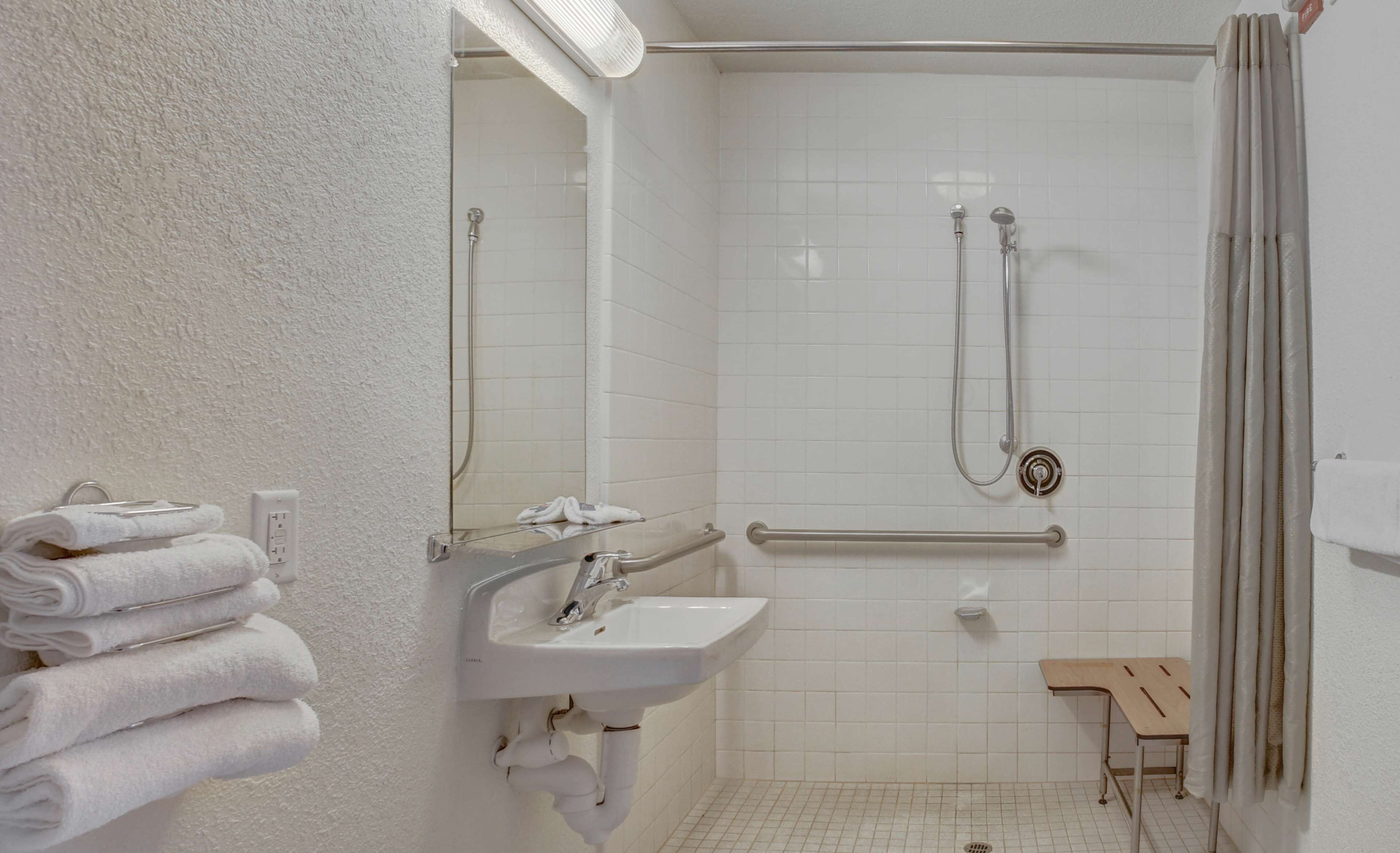A bathroom with a white sink, a mirror, a shower with a handheld showerhead, a grab bar, and a small wooden bench.