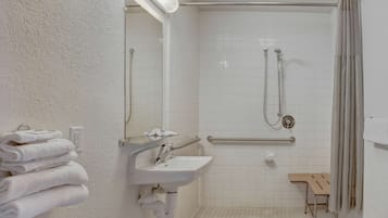 A bathroom with a white sink, a mirror, a shower with a handheld showerhead, a grab bar, and a small wooden bench.