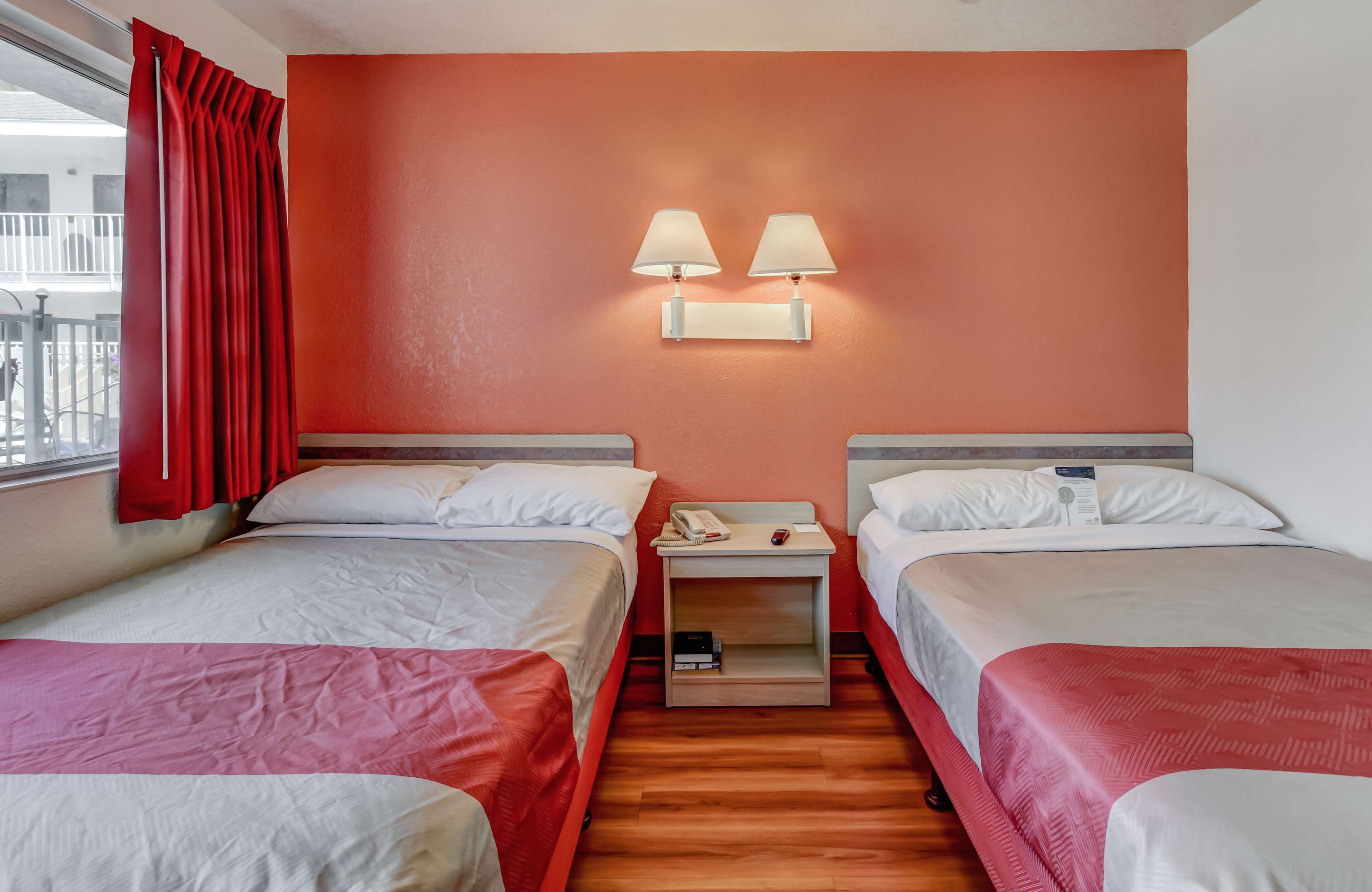 A hotel room with two beds, a nightstand with a phone and a book, and a red curtain.