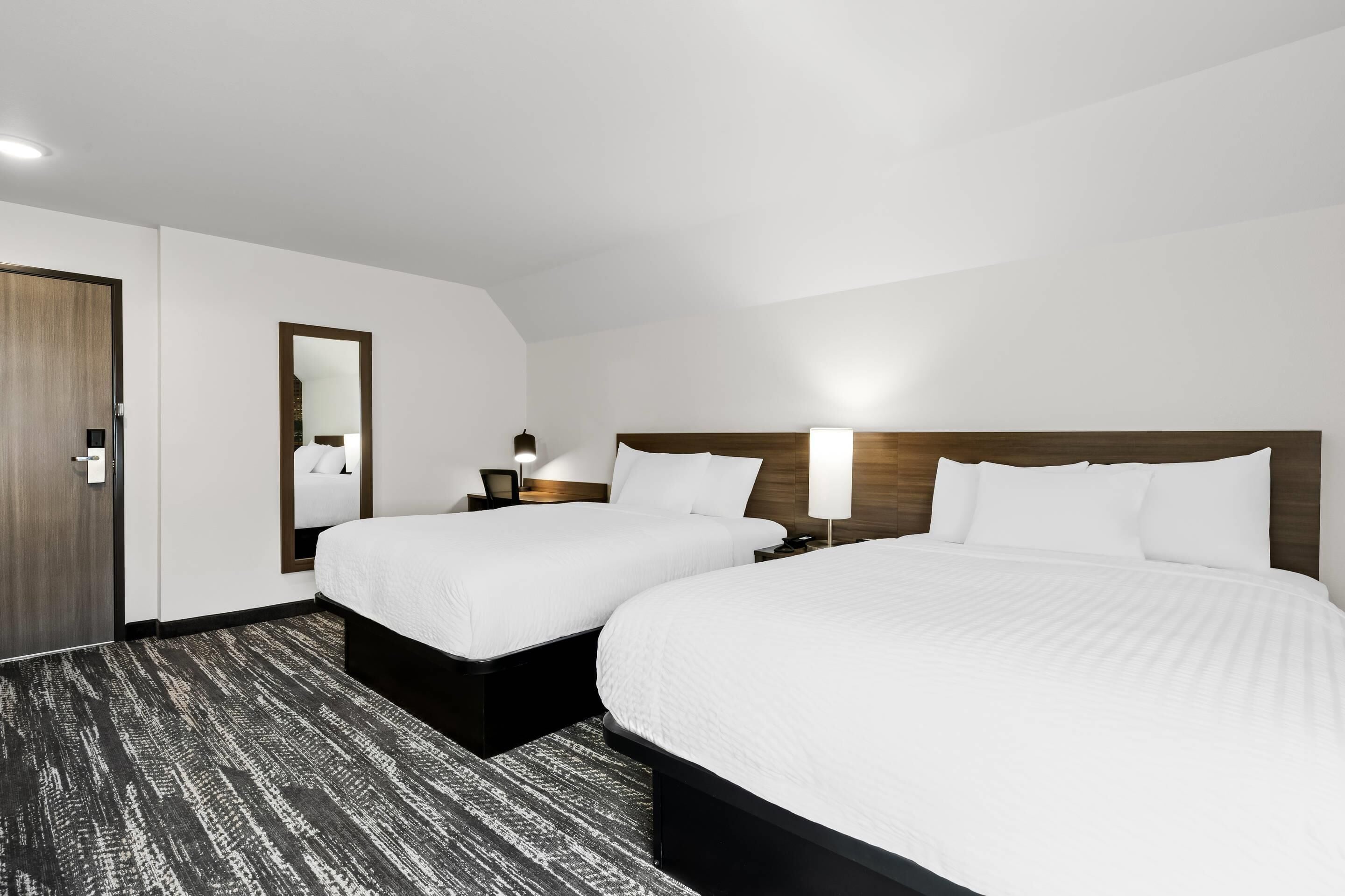 Habitación de hotel con dos camas, cabecera de madera, mesita de noche con lámpara, espejo y puerta.