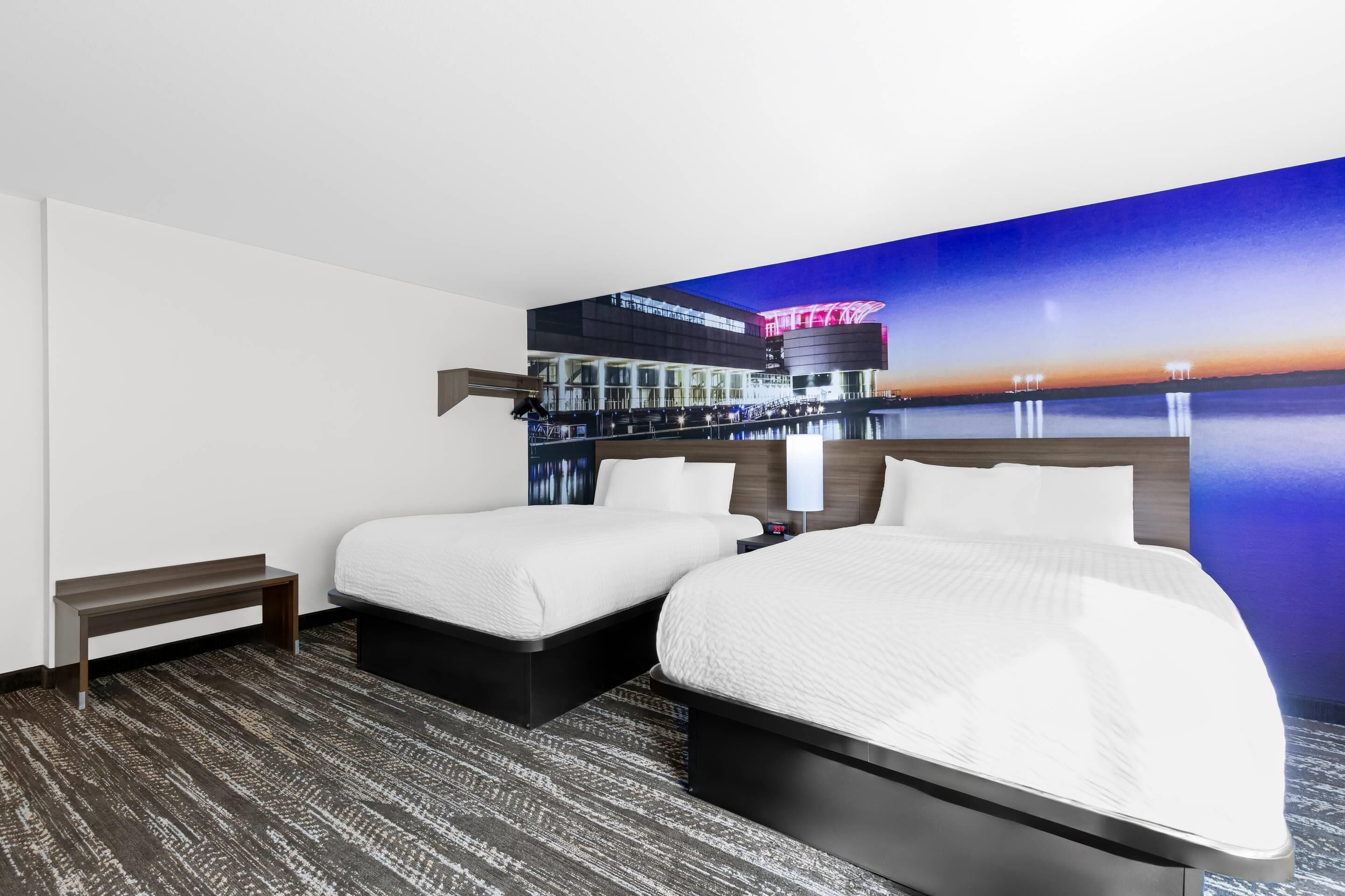 Une chambre d’hôtel avec deux lits, un banc et une grande fresque murale représentant un paysage urbain de nuit.