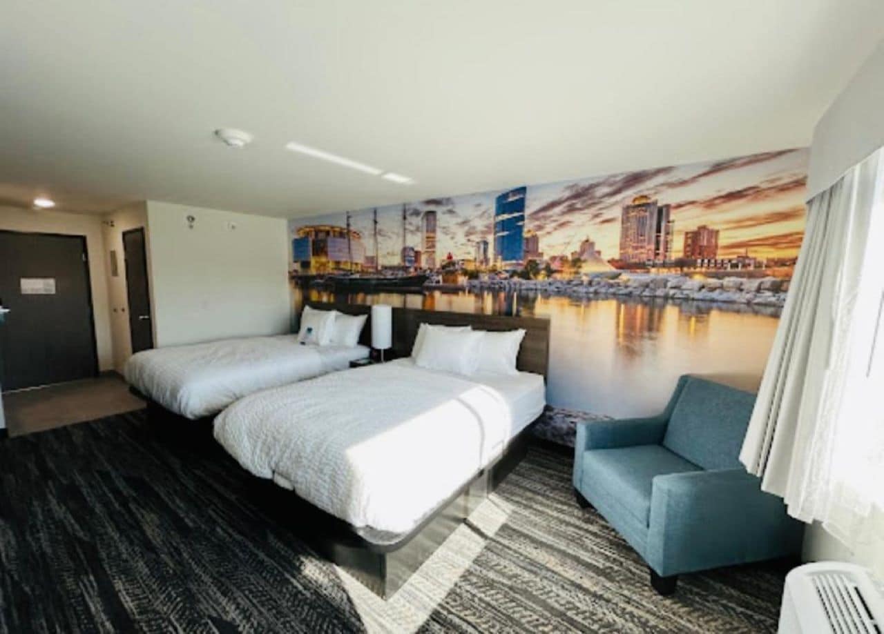 A hotel room with two beds, a blue armchair, and a cityscape mural on the wall.