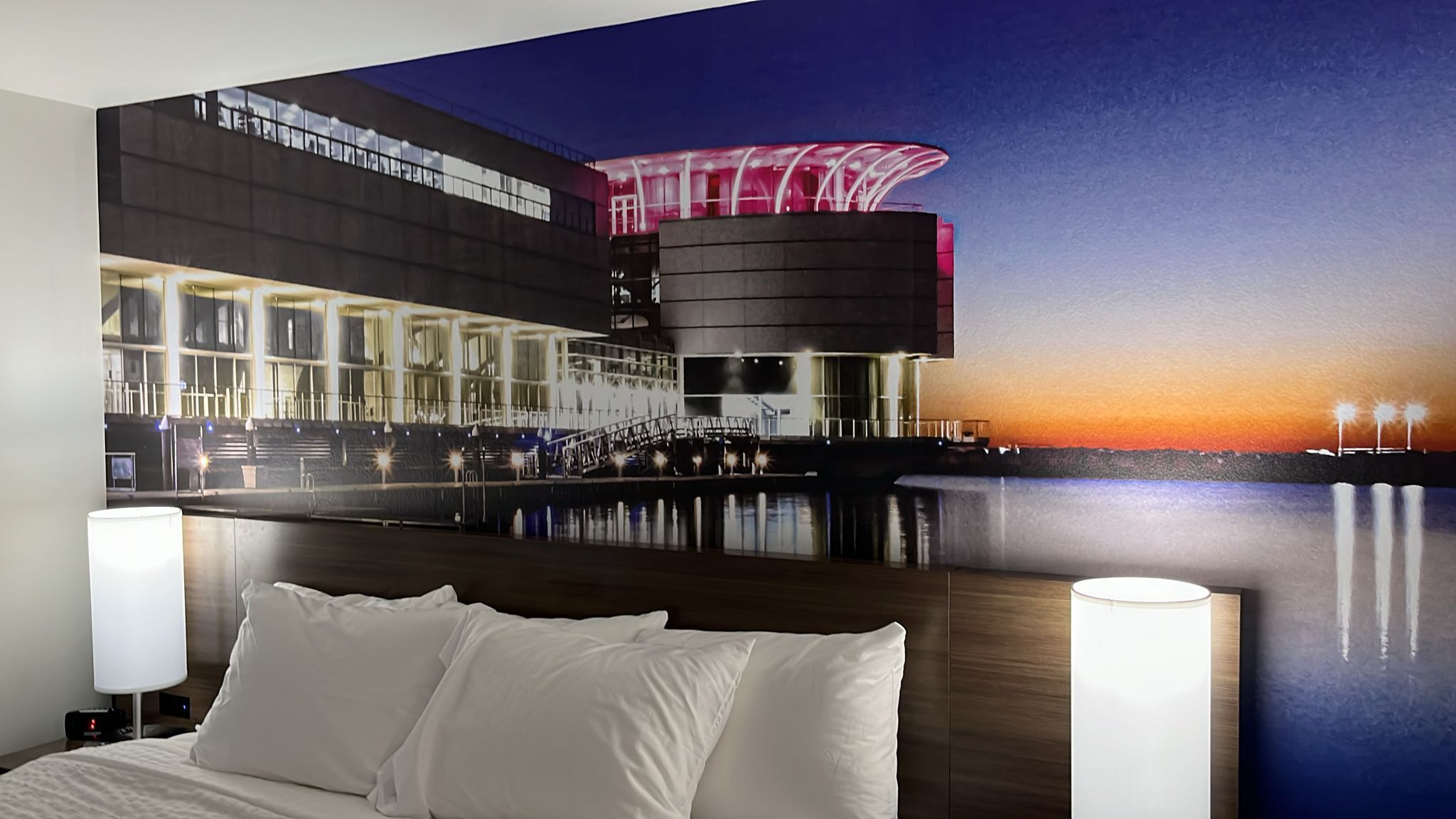 A hotel room with a large bed, a nightstand, and a wall mural of a building at sunset.