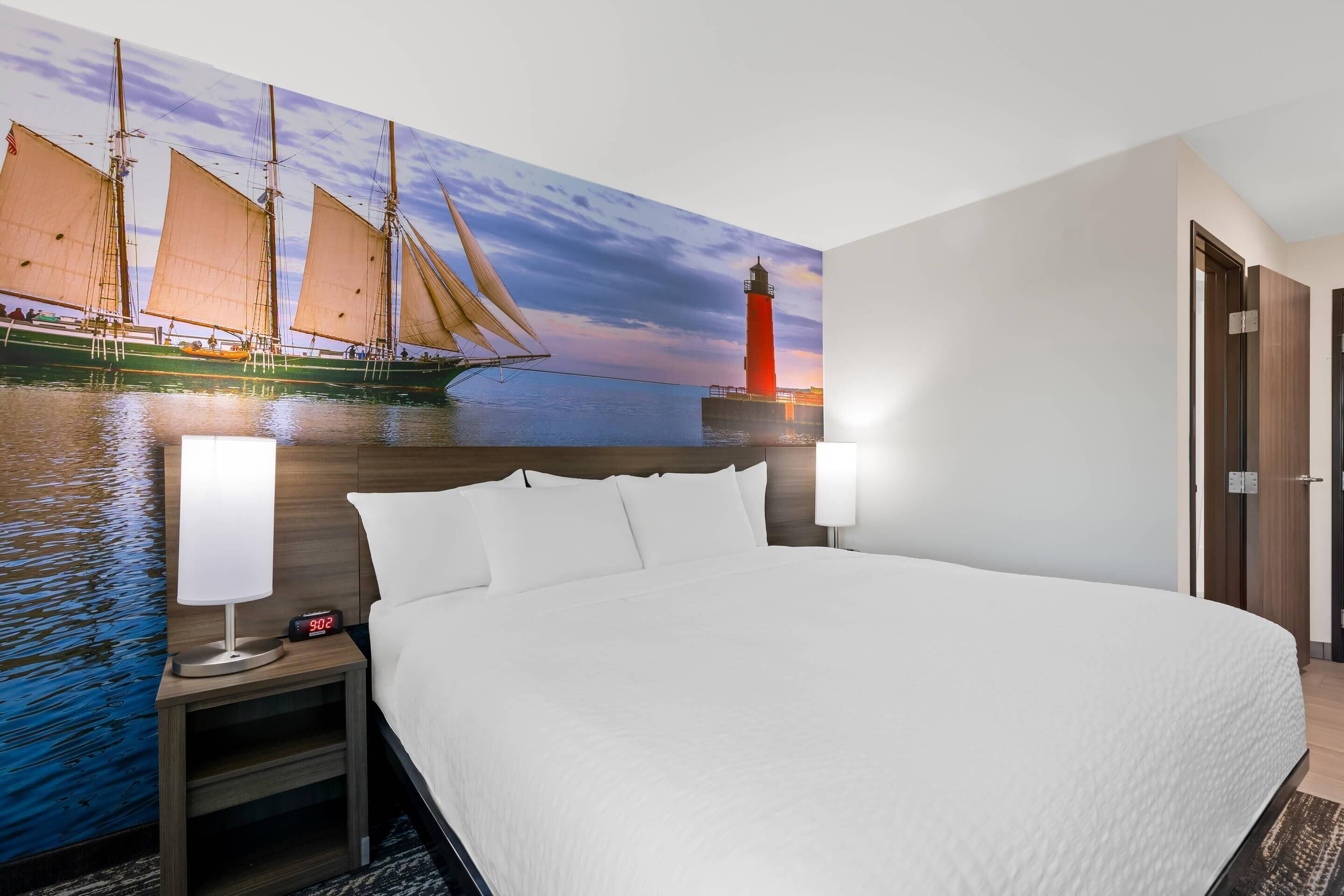 Habitación de hotel con una cama grande, dos mesitas de noche con lámparas, un tocador y un mural en la pared con un velero y un faro, además de una puerta.