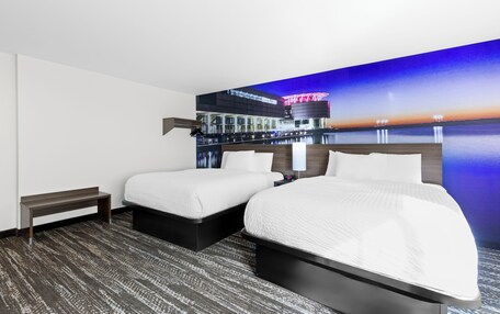 A hotel room with two beds, a bench, and a large wall mural of a cityscape at sunset.