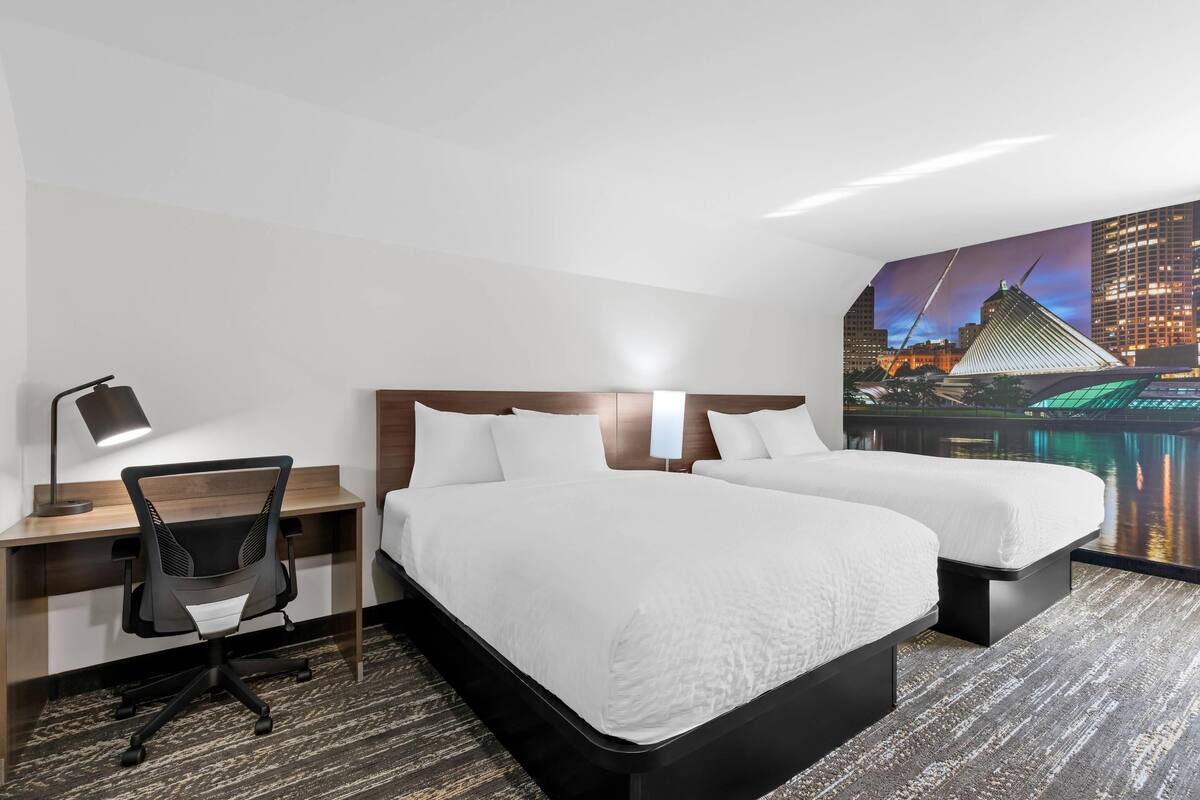 Een hotelkamer met twee bedden, een bureau, een stoel en een muurschildering van een stadsgezicht.