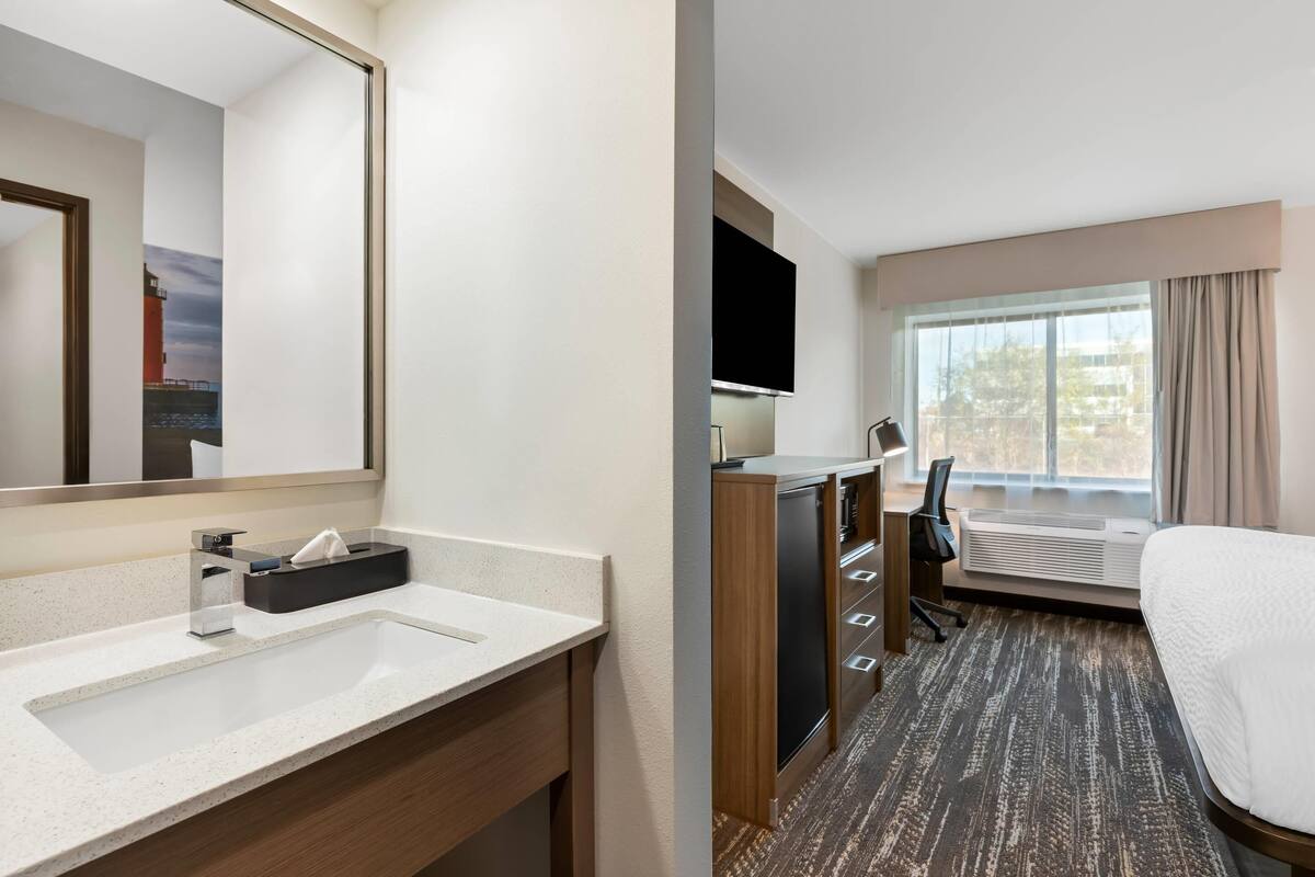Hotelkamer met een groot bed, een bureau met stoel, een televisie, een raam met gordijnen en een badkamer met wastafel en spiegel.