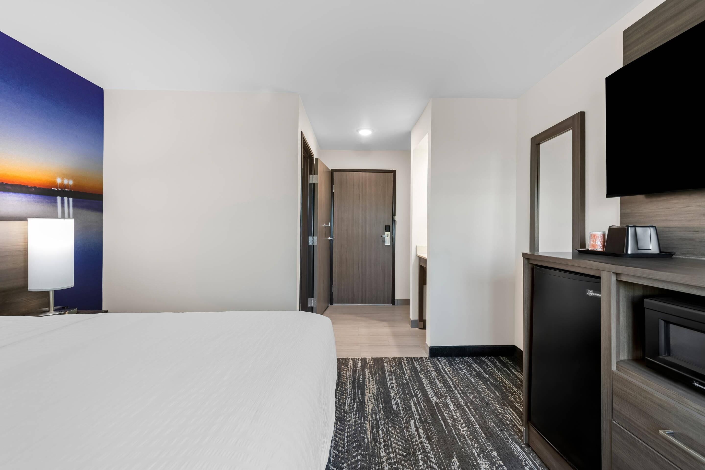 Een hotelkamer met een bed, een bureau met een flatscreen televisie, een kleine koelkast en een schilderij van een zonsondergang aan de muur.