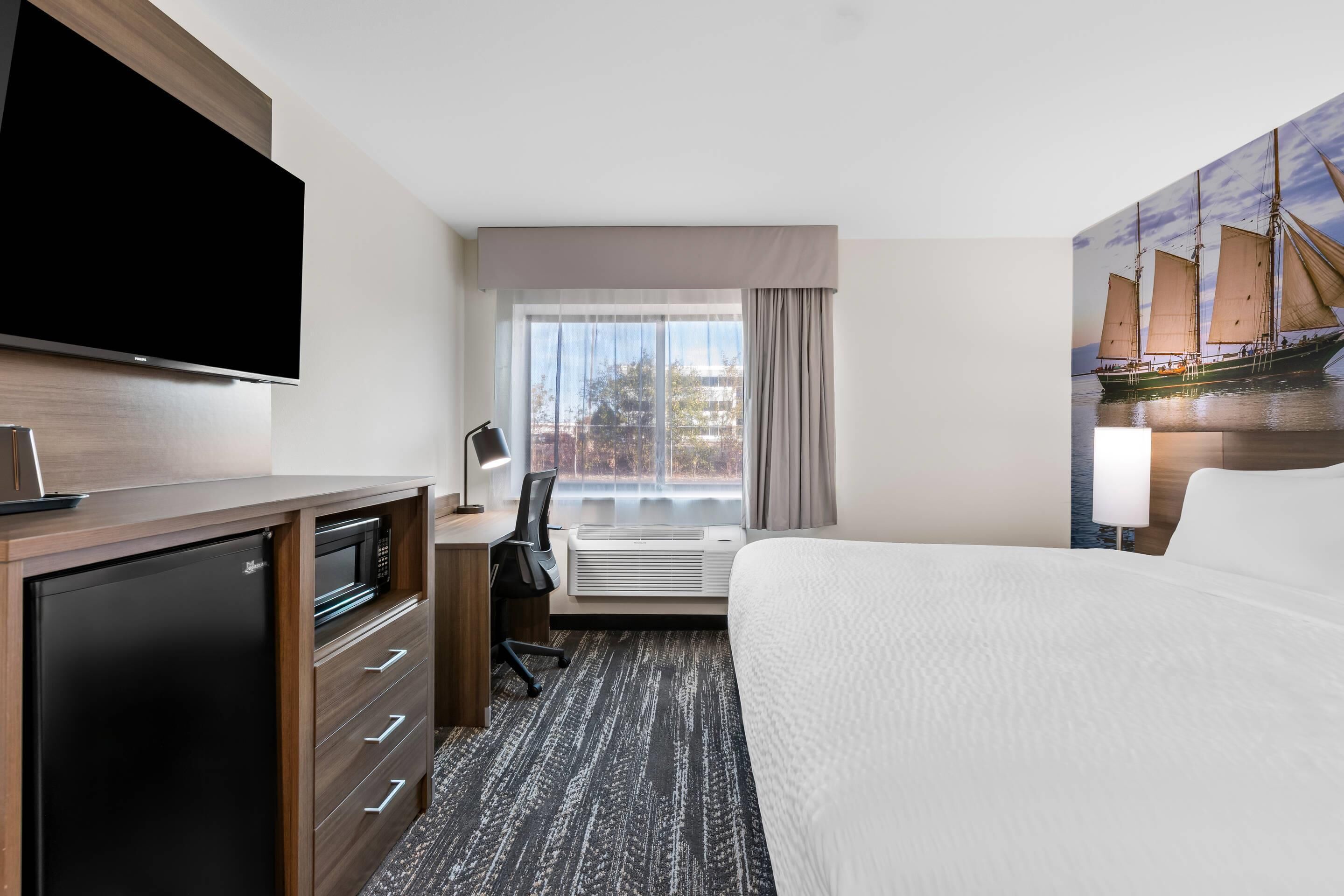 Une chambre d’hôtel comprenant un lit, un bureau avec une chaise, un téléviseur à écran plat, un mini-réfrigérateur et une grande fenêtre avec des rideaux.