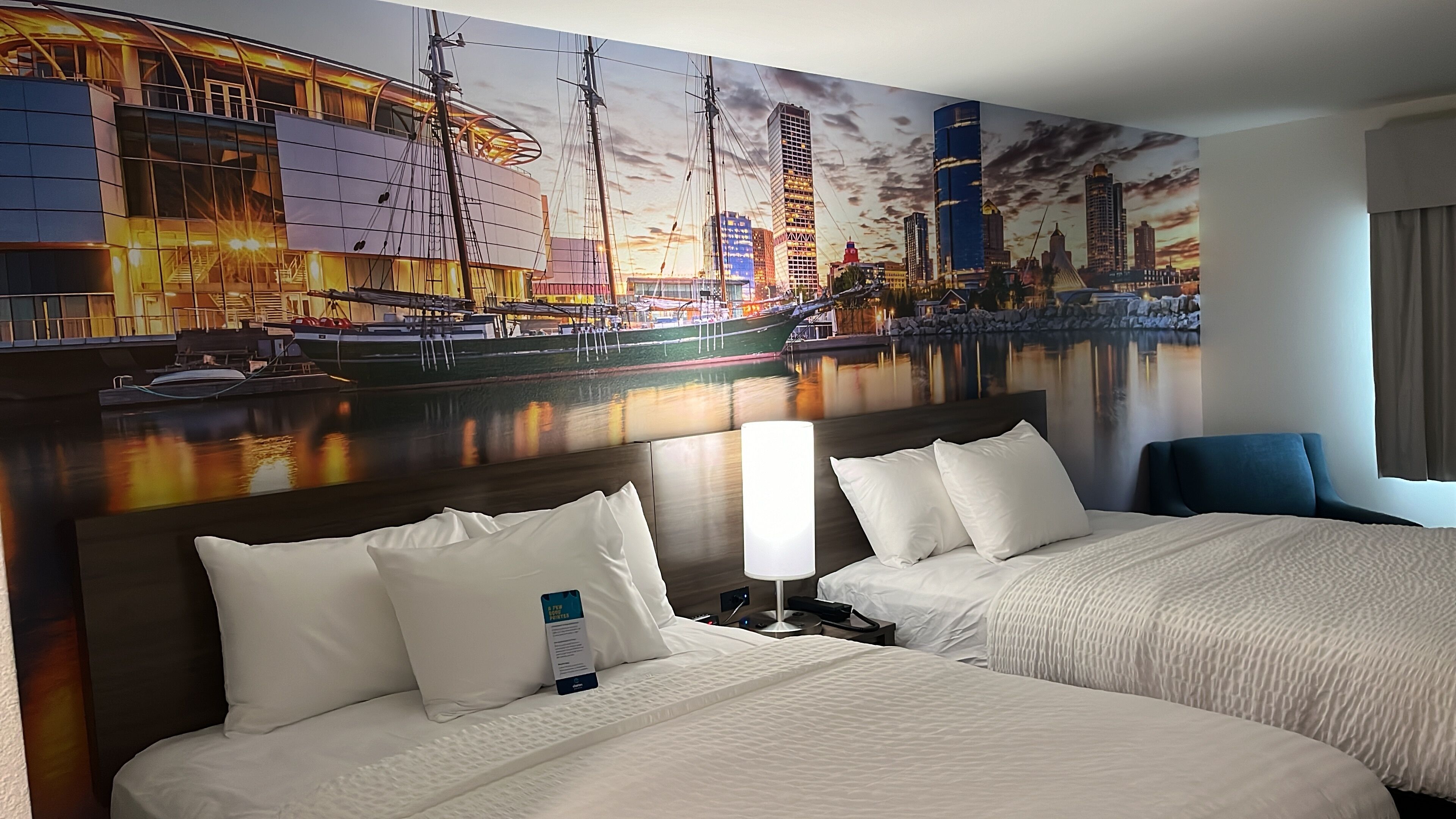 Habitación de hotel con dos camas, un mural grande con un paisaje urbano y vista al puerto.