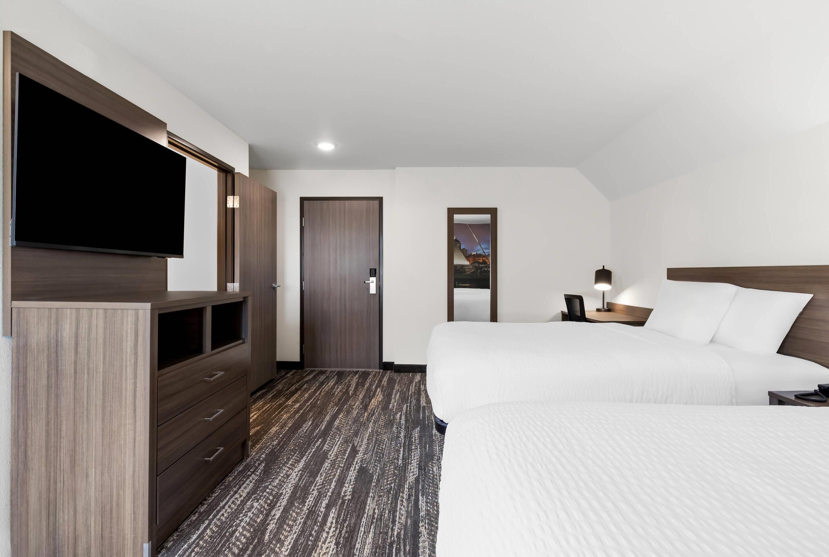 Habitación de hotel con un televisor de pantalla plana grande, un armario de madera, una cama con ropa blanca, una mesita de noche con lámpara y una puerta con espejo.
