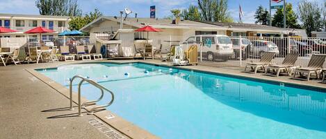 A swimming pool with lounge chairs and umbrellas, surrounded by a fenced area with a building in the background.
