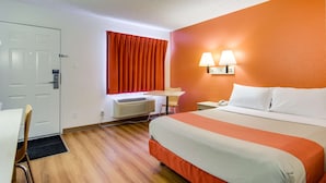 Una habitación de hotel con cama, escritorio, silla, lámpara de escritorio y ventana con cortinas.