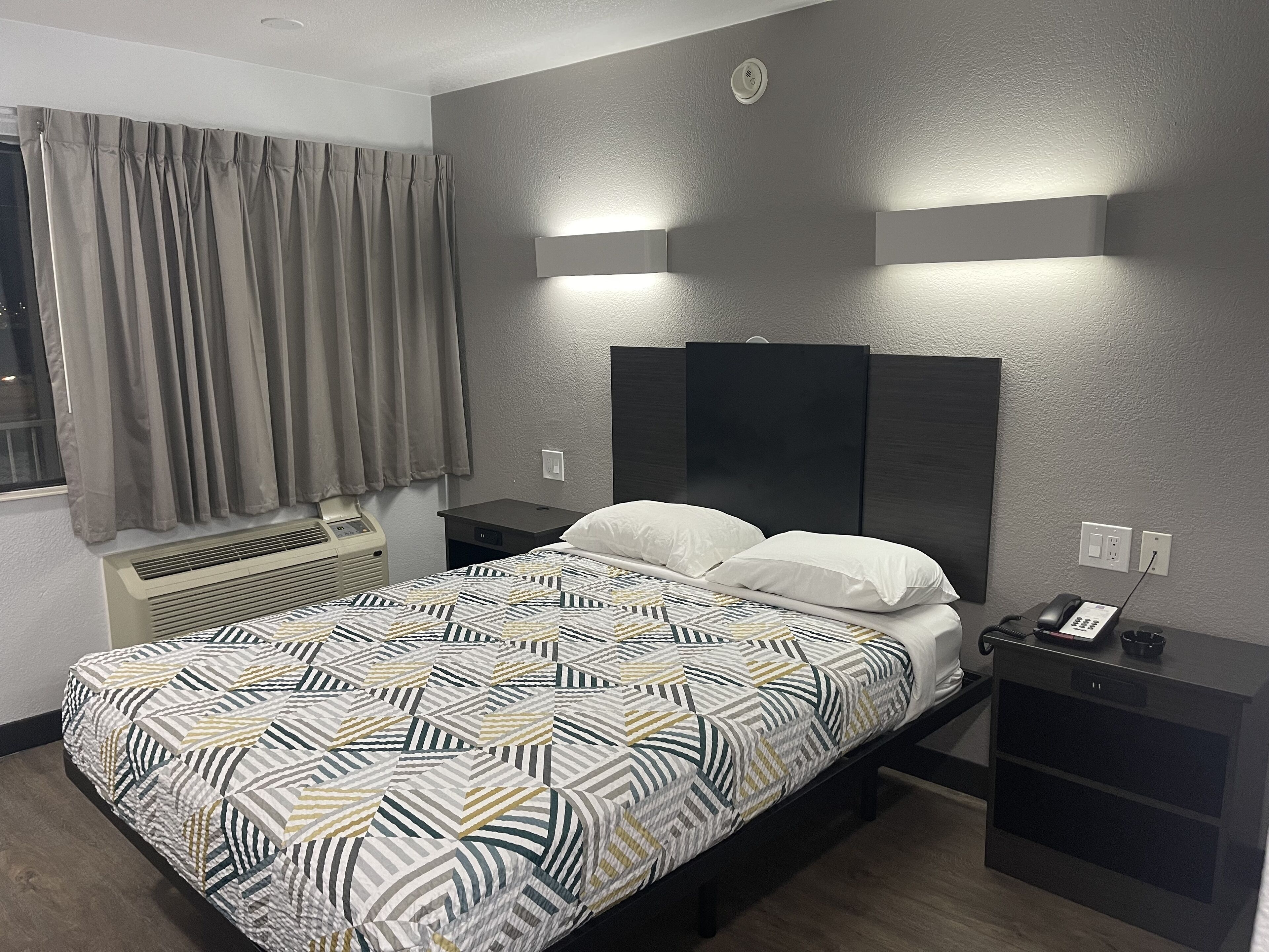 A modern hotel room with a bed, bedside tables, a heater, a phone, and a window with curtains.