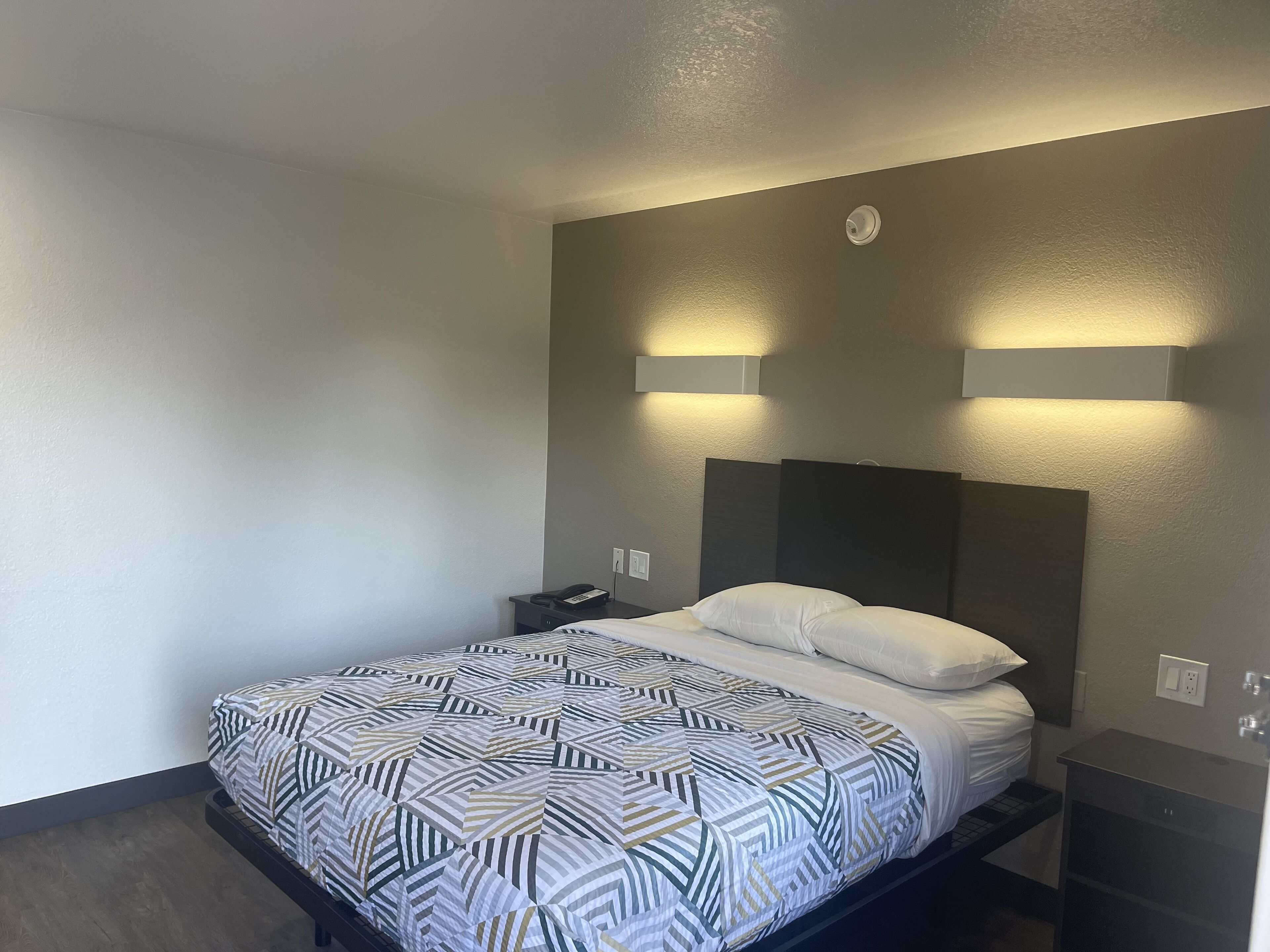 A hotel room with a bed, bedside table, and a wall-mounted light fixture.