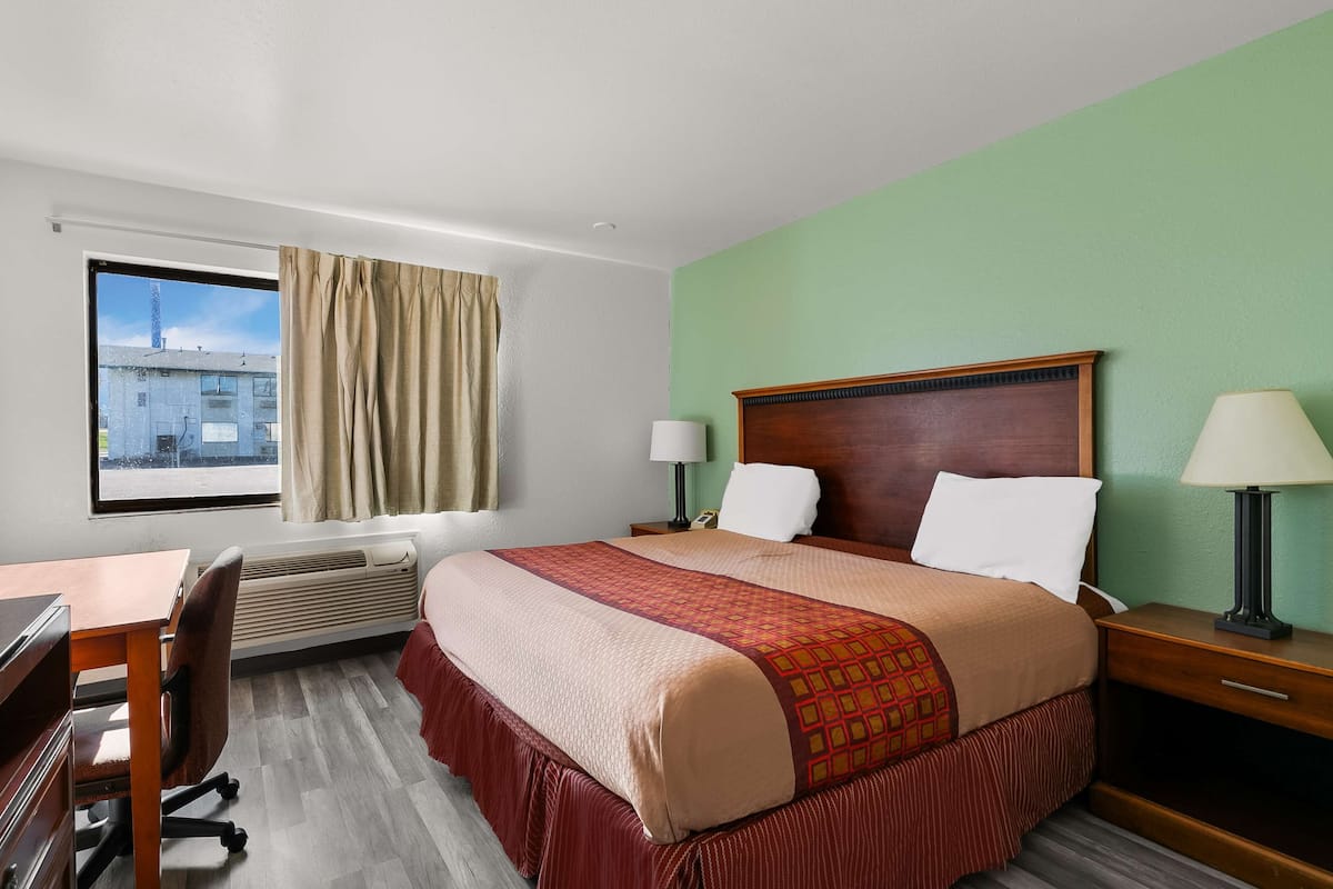 A hotel room with a large bed, two bedside tables with lamps, a desk with a chair, a window with curtains, and an air conditioning unit.
