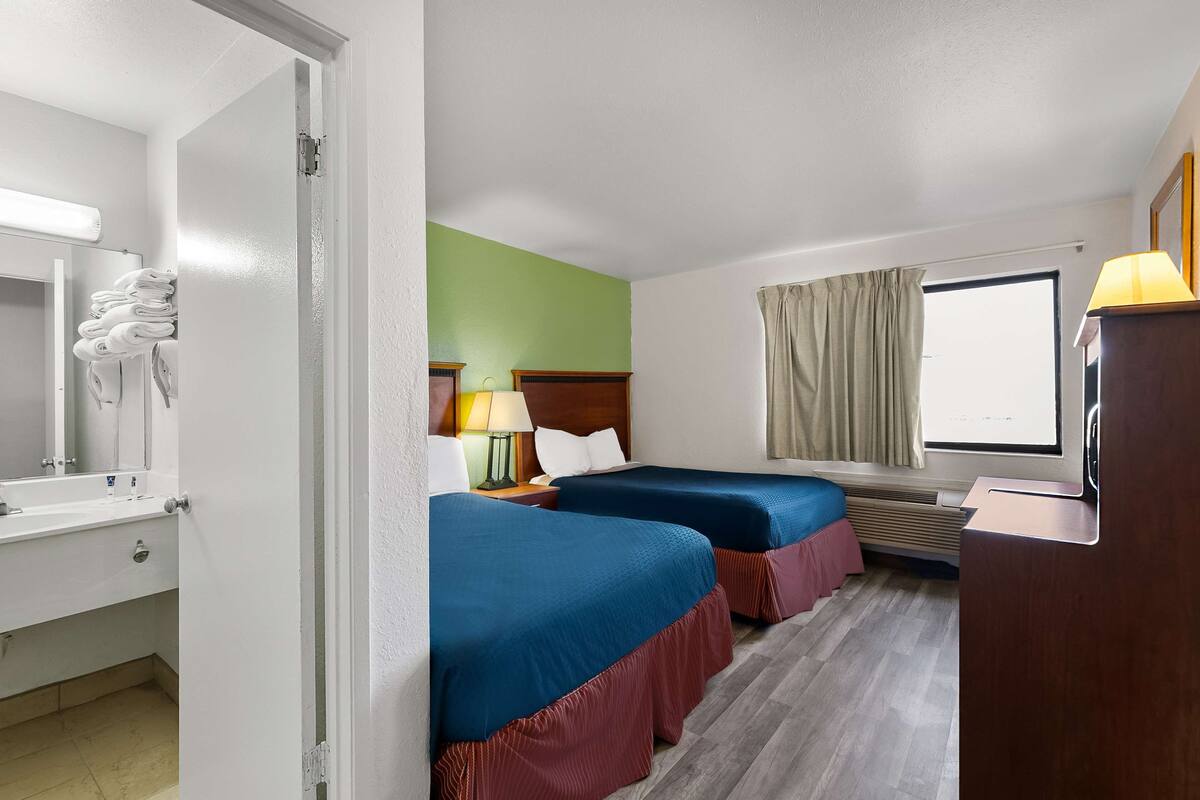 A hotel room with two beds, a bathroom with a mirror and towel rack, a desk with a lamp, and a window with curtains.