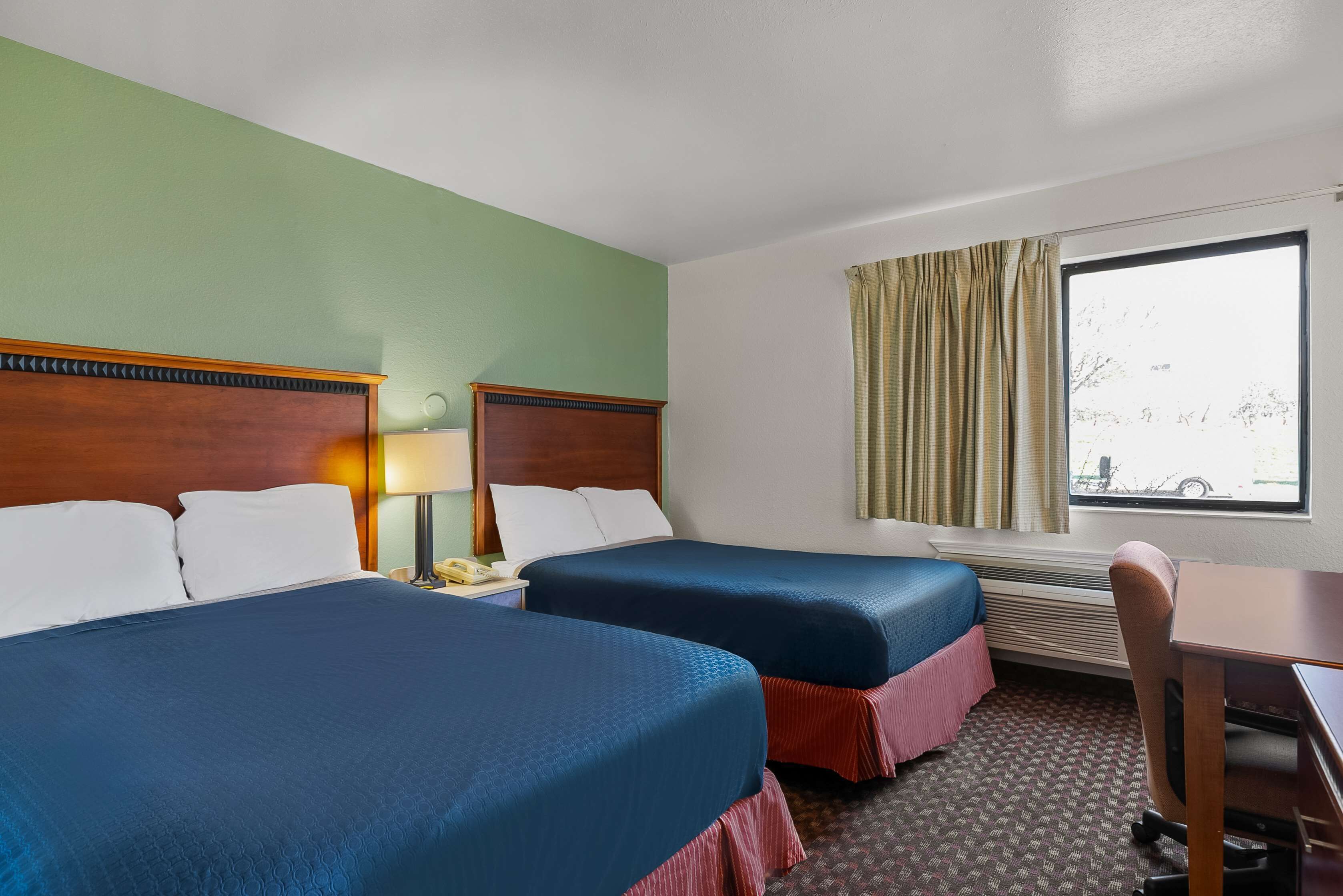 A hotel room with two beds, a desk, a chair, a lamp, and a window with curtains.