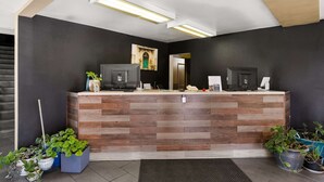 Een receptiebalie met een houten afwerking, twee zwarte monitoren, een telefoon en een koffieapparaat. Aan weerszijden van de balie staan plantenpotten.