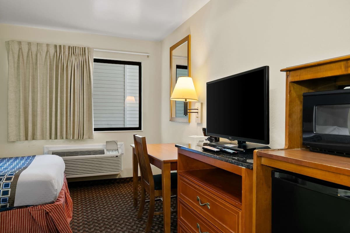 A hotel room with a bed, a desk with a flat-screen TV, a chair, a dresser, and a window with curtains.