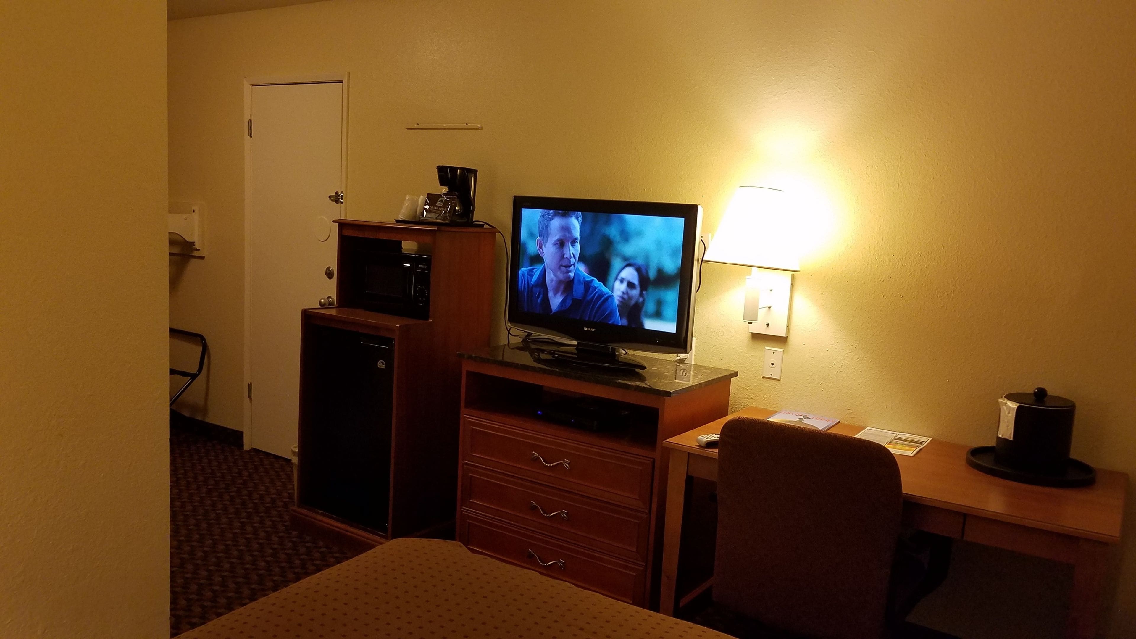 A hotel room with a television, a desk, a chair, a lamp, and a coffee maker.