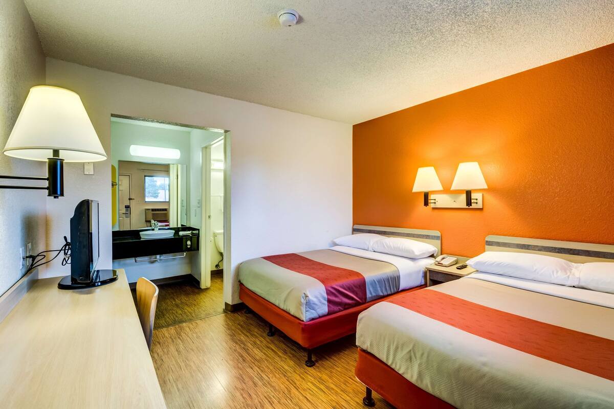 A hotel room with two beds, a desk, a chair, a TV, and a bathroom visible through an open door.