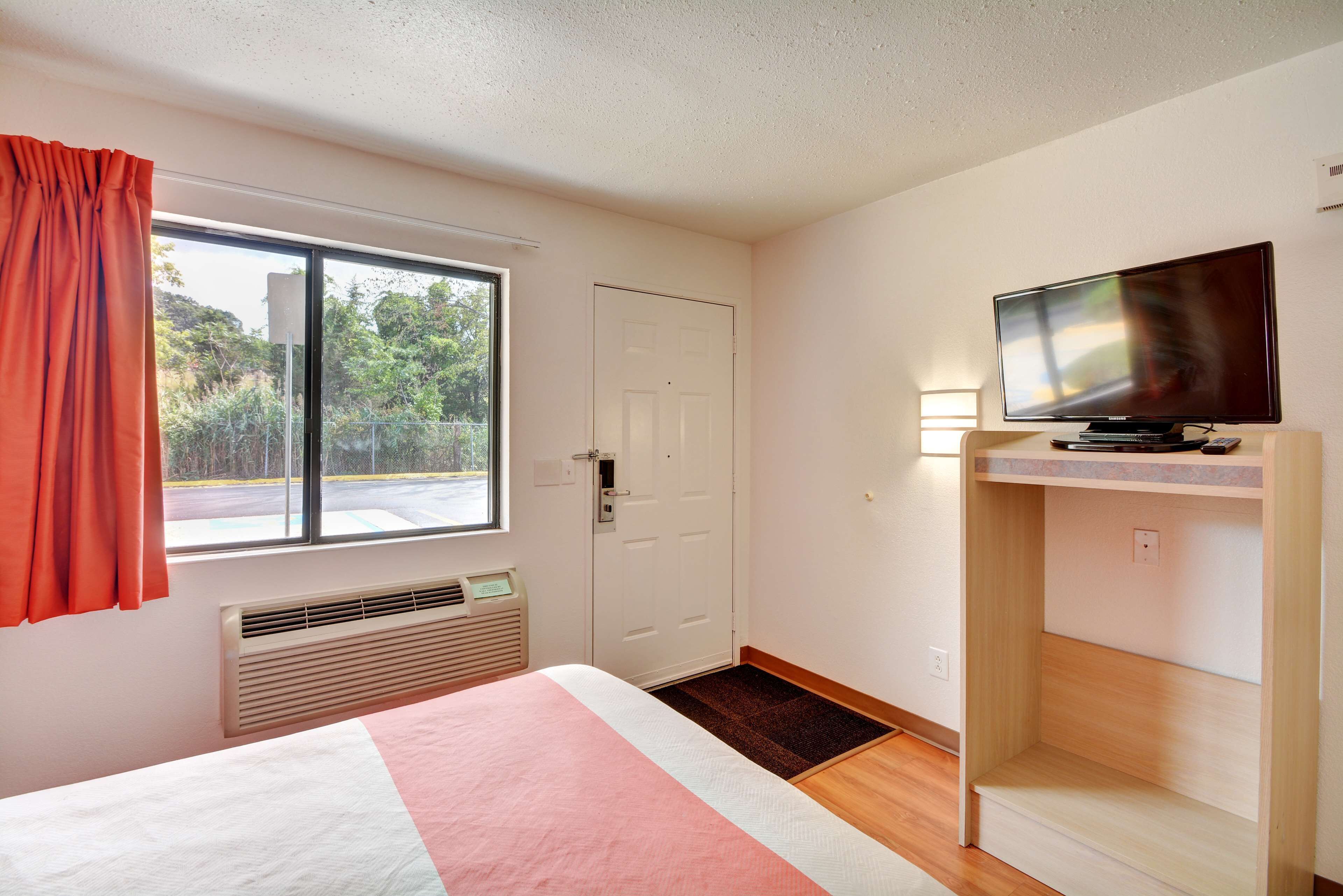 A hotel room with a bed, a television, a window with curtains, a door, and a view of the outdoors.