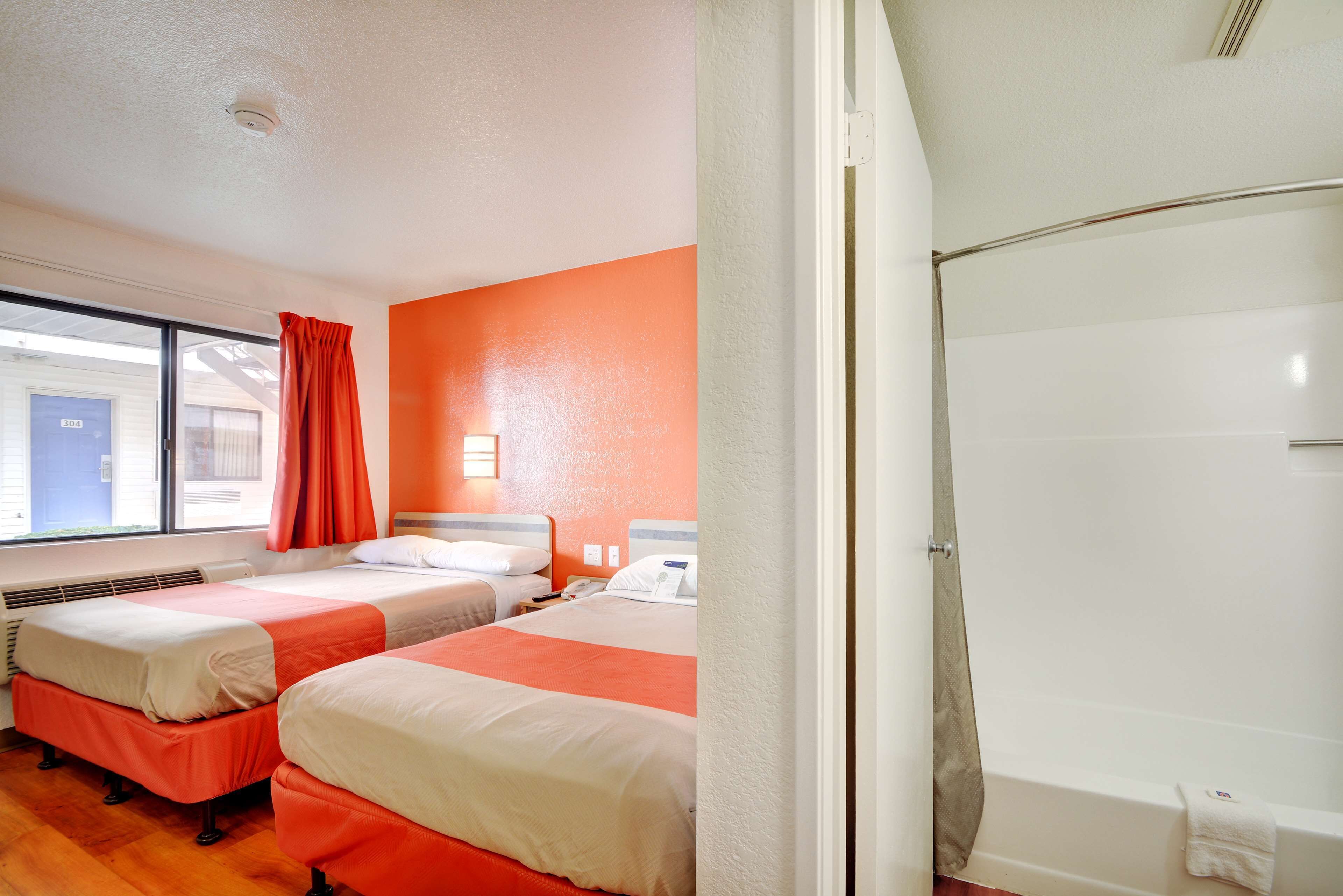 A hotel room with two beds, a window with curtains, a white bathtub, and a visible door.