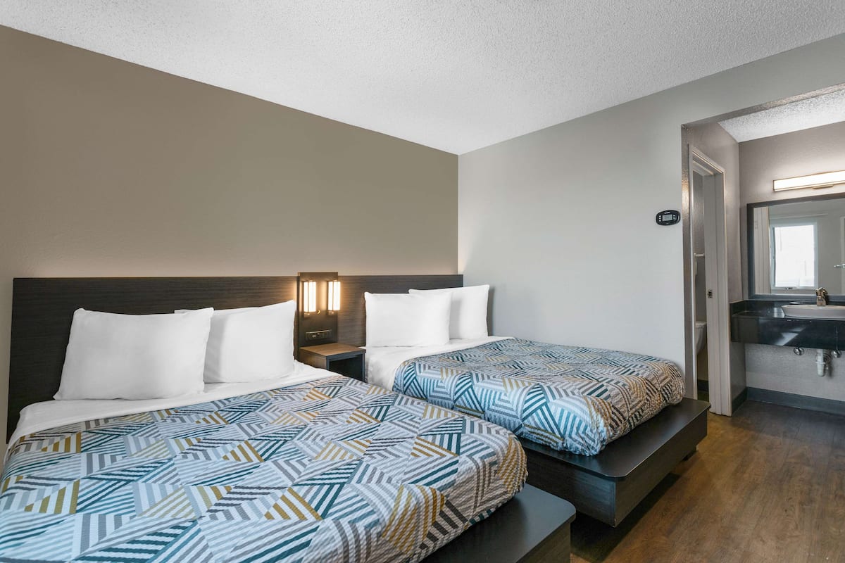 A hotel room with two beds, a nightstand, a wall-mounted light, and a bathroom with a mirror and sink.