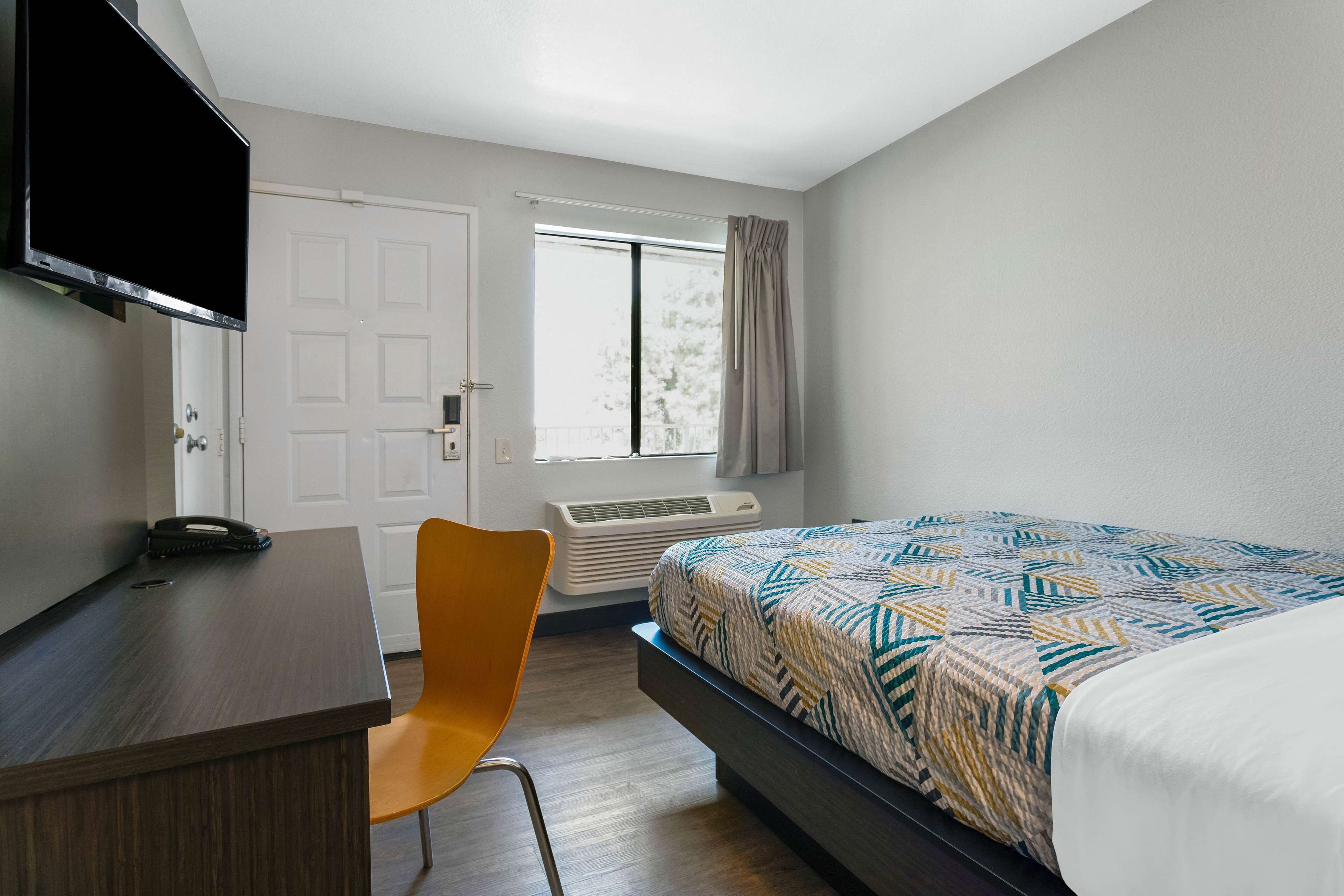 A bedroom with a bed, a desk, a chair, a TV, a window, and an air conditioning unit.