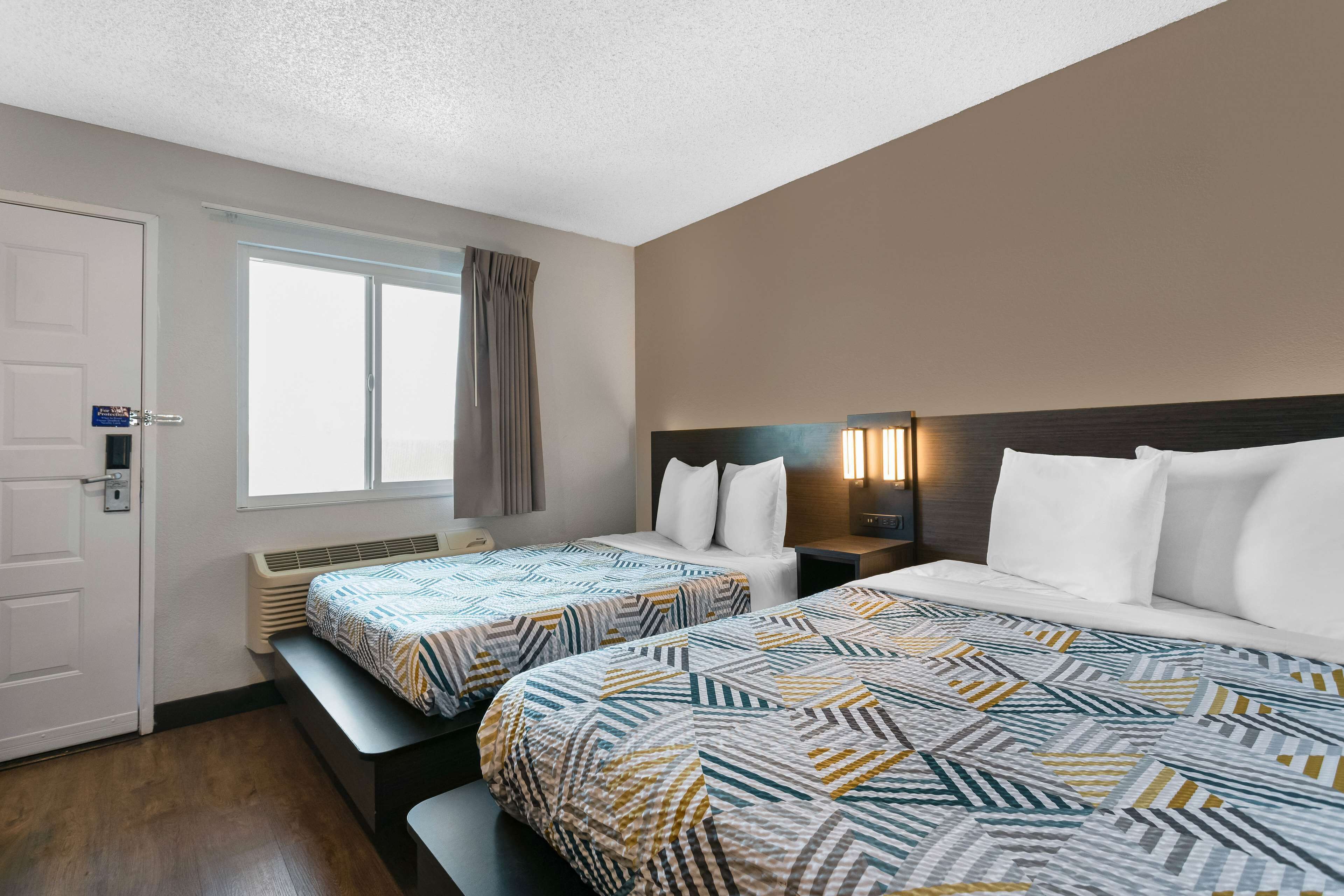 A hotel room with two beds, a window with curtains, a bedside table, and a wall-mounted lamp.