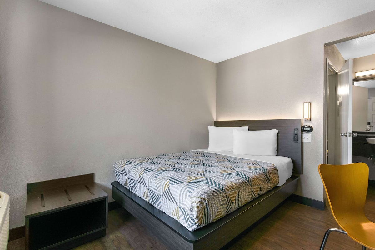 A hotel room with a bed, a chair, a bedside table, and a small storage unit.