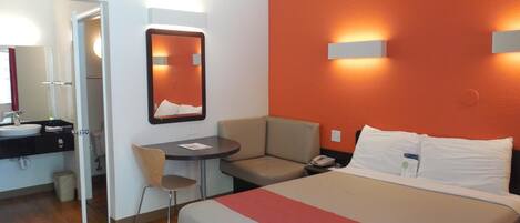 A hotel room with a bed, a desk, a chair, a mirror, and a bathroom.