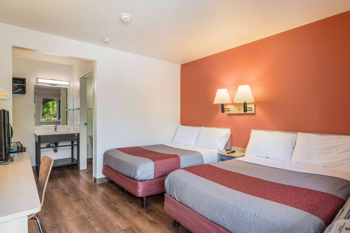 A hotel room with two beds, a desk, a mirror, and a bathroom visible through an open door.