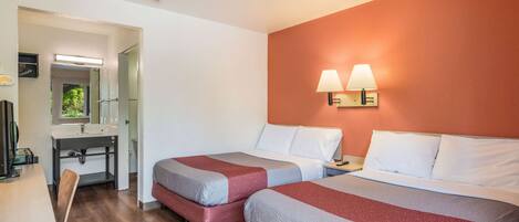 Quarto de hotel com duas camas, uma escrivaninha, um espelho e um banheiro visível através de uma porta aberta.