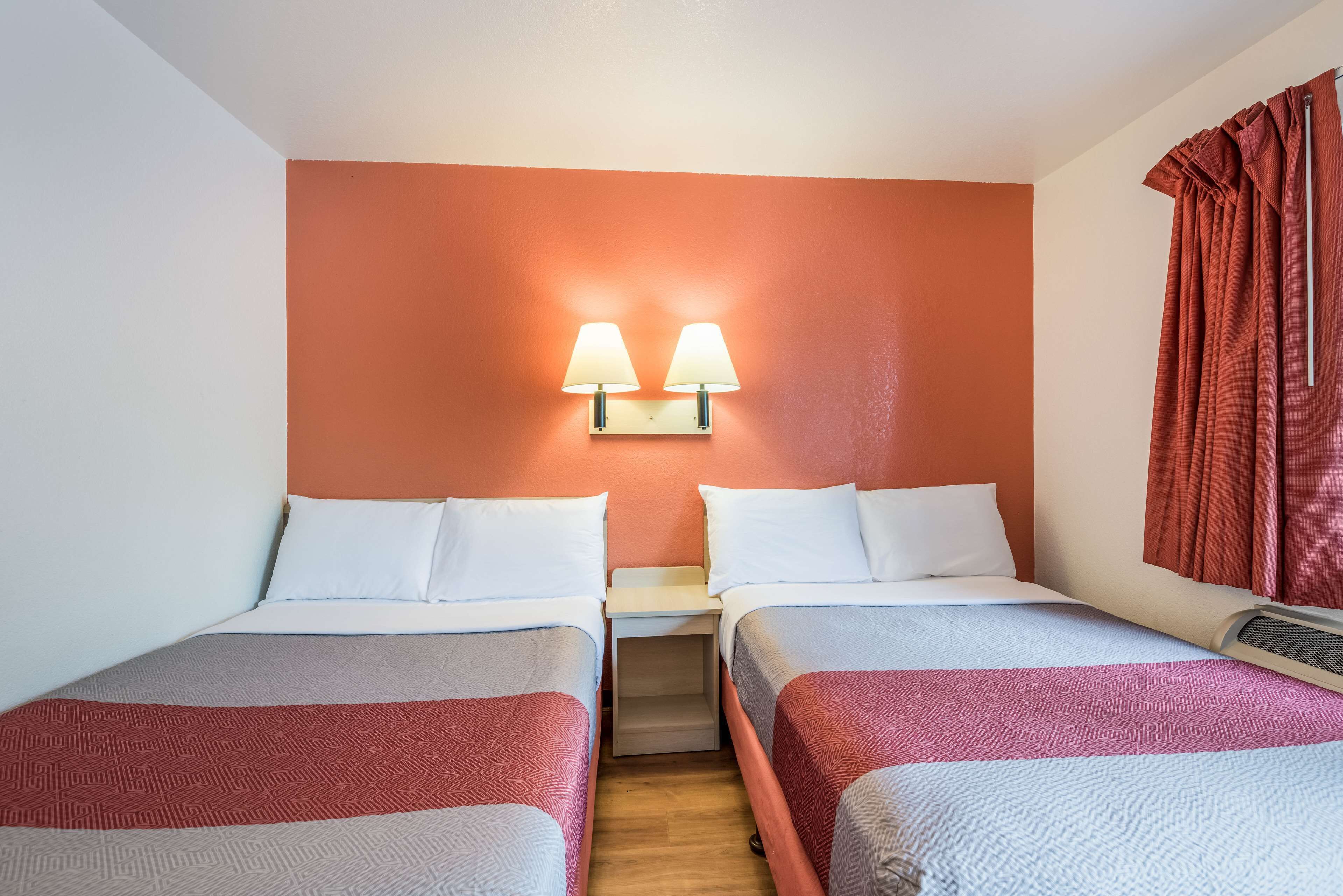 A hotel room with two beds, a nightstand, a wall-mounted lamp, and a window with curtains.