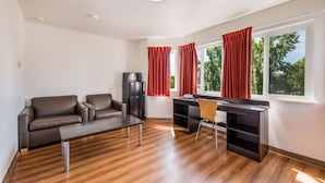 A modern living room with a brown leather sofa, a wooden coffee table, a dark desk with a chair, and a black refrigerator. - Motel 6 Rohnert Park, CA (Rohnert Park)