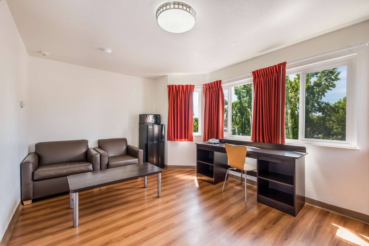 A modern living room with a brown leather sofa, a wooden coffee table, a dark desk with a chair, and a black refrigerator.