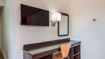 A hotel room with a flat-screen TV mounted on the wall, a bedside lamp, a wooden dresser, and a chair.