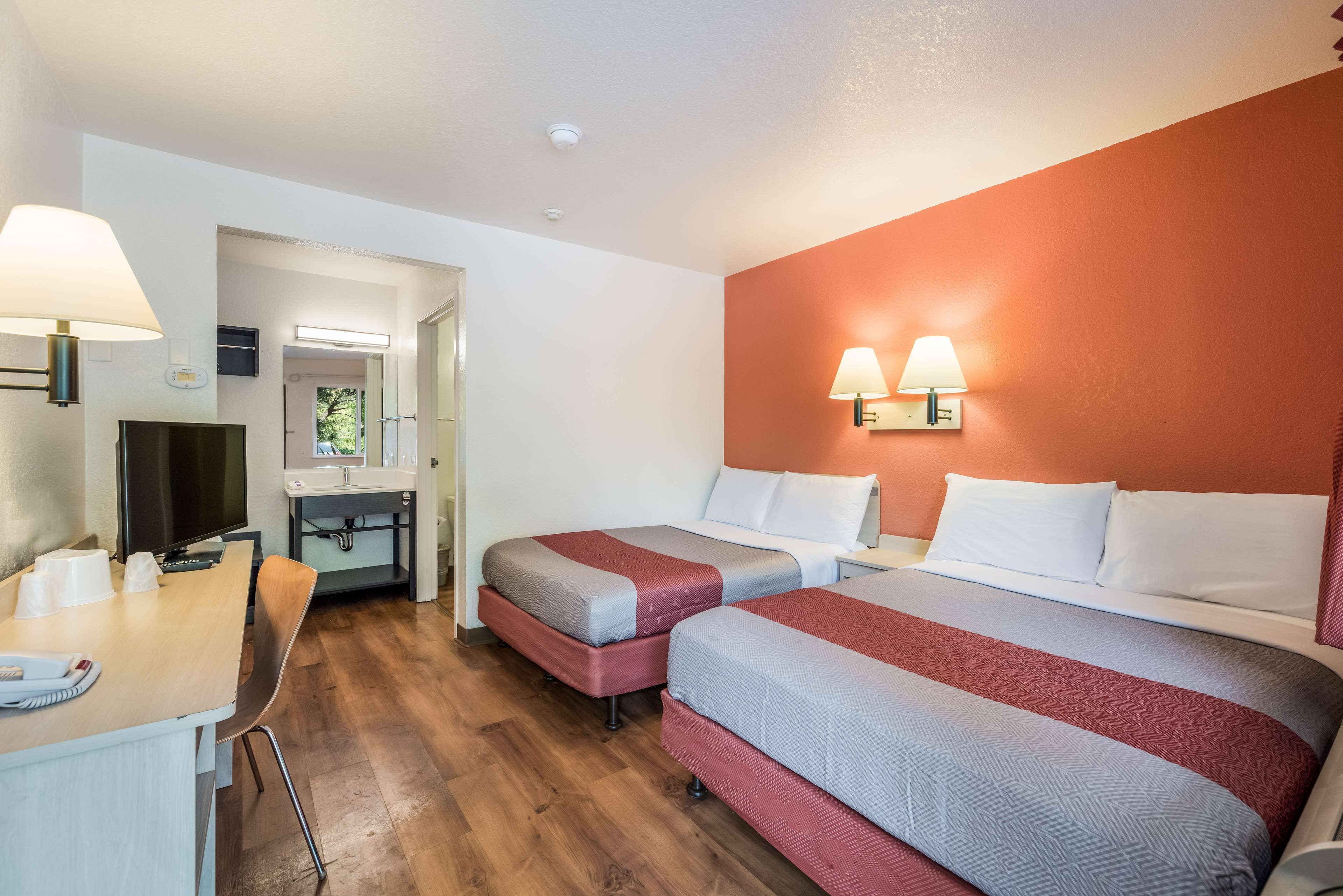 A hotel room with two beds, a desk with a lamp, a television, and a bathroom visible through an open door.